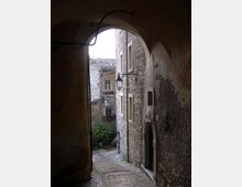 Passaggio ad arco in pietra che si apre su una stretta viuzza in discesa lastricata, tra edifici in pietra con finestre piccole e portale ad arco. Lanterne in ferro battuto sporgono dalle pareti e si intravedono tetti in coppi e alcune piante in fondo al vicolo.