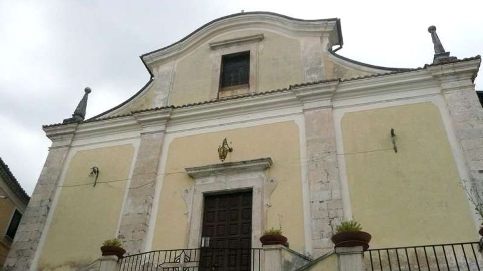 Facciata di una chiesa in stile semplice con intonaco giallo e cornici in pietra chiara, timpano curvilineo e una finestra centrale superiore. In basso si vede l’ingresso con portale scuro, una scalinata con ringhiera in ferro e vasi di piante su pilastri, sotto un cielo nuvoloso.