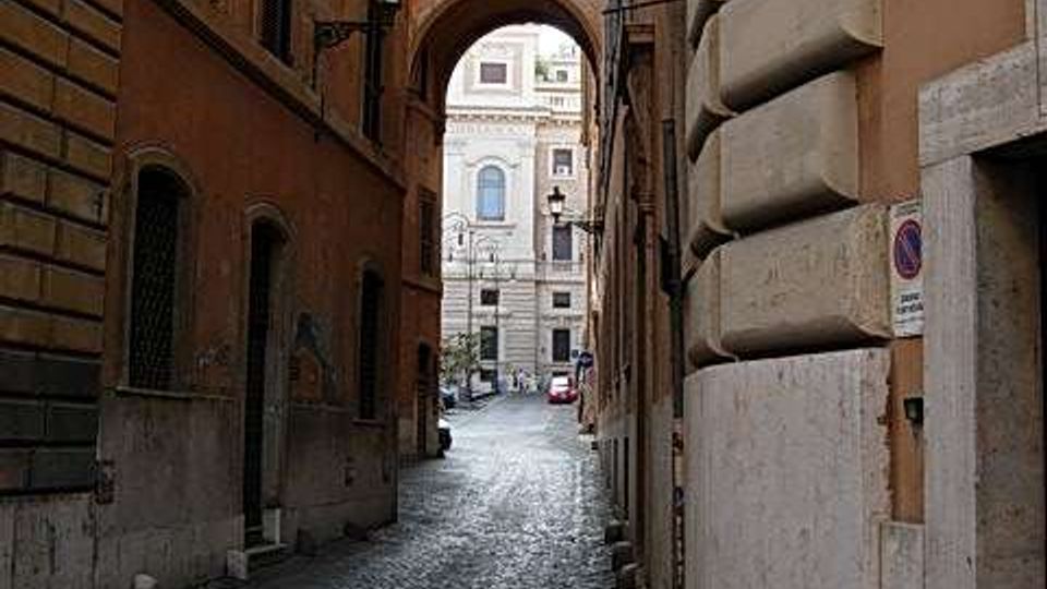 Vicolo urbano stretto con pavimentazione in sampietrini, fiancheggiato da alti edifici in muratura con finestre e un lampione a parete. In fondo si vede un arco che collega i palazzi e apre su una strada più ampia con altri edifici.