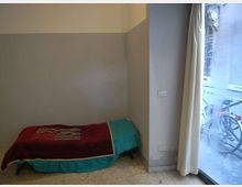 Piccola camera con letto singolo basso addossato alla parete, pareti bicolore grigio e bianco e pavimento in graniglia. A destra una grande finestra o porta-finestra con tende chiare affaccia sulla strada esterna.