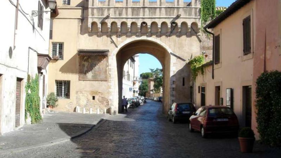 Strada urbana in ciottoli che attraversa un grande arco in muratura con merlature e piccole arcate decorative, inserito tra edifici storici. Ai lati della via si vedono facciate intonacate con finestre e alcune auto parcheggiate.