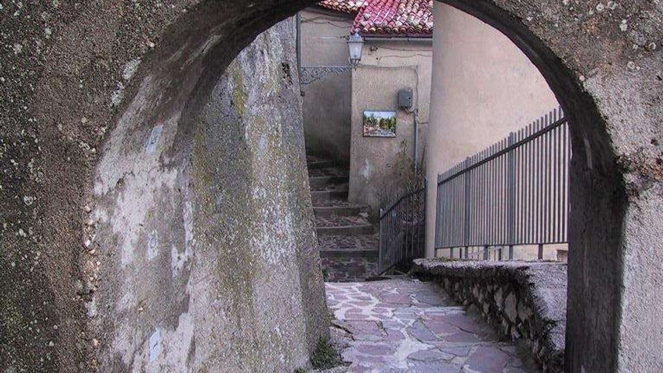 Arco in pietra scura che incornicia un vicolo stretto in discesa con pavimentazione irregolare in lastre di pietra, tra muri intonacati e una ringhiera metallica. In fondo si vedono scale, un tetto in coppi rossi e una targhetta con scritta sopra l’arco.