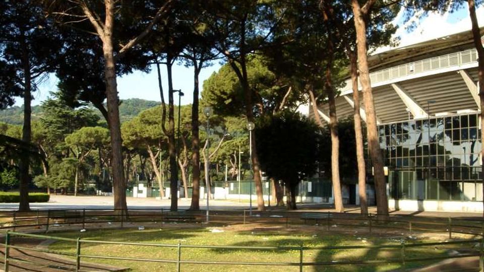 Parco con alti pini e aiuole erbose delimitate da ringhiere basse e panchine lungo vialetti. Sul lato destro si vede un grande edificio moderno con facciata vetrata e ampia struttura sporgente.