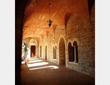 Portico in stile storico con volta in mattoni e pareti in pietra, illuminato dalla luce laterale che crea ombre sul pavimento in cotto. Lungo il lato destro si susseguono arcate con colonne e nicchie, con lanterne sospese e alcune piante in vaso vicino all’ingresso sul fondo.