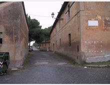 Strada stretta tra due lunghi edifici in intonaco marrone dall’aspetto rurale, con piccole finestre e cavi aerei, illuminata da un lampione sospeso. La carreggiata è in parte acciottolata e conduce verso alberi alti e una strada principale sullo sfondo.