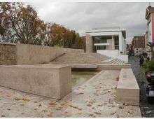 Spazio urbano con ampia scalinata e piattaforme in pietra chiara che circondano una vasca d’acqua bassa. Sullo sfondo si vede un edificio moderno bianco con grandi vetrate, affiancato da alberi e da una strada cittadina.