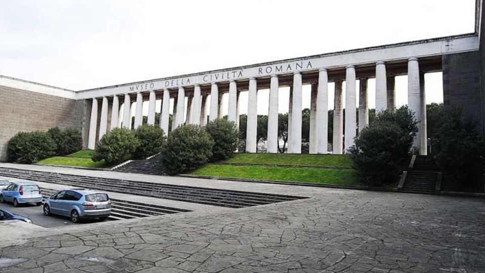 Facciata del Museo della Civiltà Romana con lunga colonnata di pilastri bianchi e scritta in sommità, affacciata su una piazza pavimentata con ampie scalinate. Davanti si vedono aiuole verdi con arbusti e un’area di parcheggio ai margini.