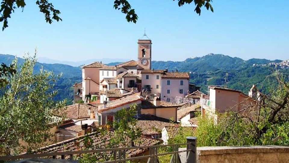 Veduta panoramica di un borgo collinare con case dai tetti in coppi e una torre campanaria con orologio al centro. In primo piano si vede una ringhiera e vegetazione, con montagne boscose sullo sfondo sotto un cielo sereno.