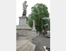 Statue in pietra su alto basamento con gradini all’ingresso di Ponte Sant’Angelo, con un’altra statua allineata lungo il parapetto. A destra si vede il cartello stradale “ponte S. Angelo”, una fila di alberi e una strada con bancarelle.