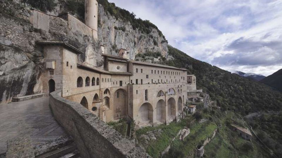 Grande complesso monastico in pietra addossato a una parete rocciosa, con lunghe facciate ad archi e un camminamento con scala e parapetto in primo piano. Sullo sfondo si vede una valle montana boscosa e cielo nuvoloso.