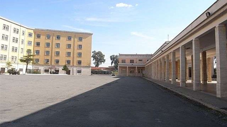 Ampia piazza asfaltata con edifici rettangolari a più piani sul lato sinistro e un lungo portico colonnato con copertura sul lato destro. Sullo sfondo si vedono altri edifici bassi e alcuni alberi sotto un cielo sereno.