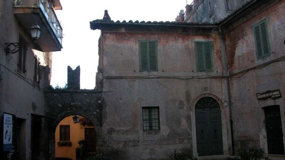 Cortile interno in un centro storico, circondato da edifici in intonaco consumato con persiane verdi e tetti in coppi. A sinistra si vede un arco in pietra che conduce a un passaggio illuminato, con porte e finestre ad arco e vasi di piante lungo il pavimento in pietra.