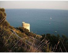 Torre costiera in pietra su un promontorio roccioso e vegetato, affacciata sul mare aperto. In lontananza si vede l’orizzonte marino con alcune barche a vela.