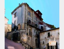 Edificio storico in pietra e intonaco su una strada in salita, con balconi in ferro battuto e finestre irregolari. Alla base si vedono muri di contenimento e un passaggio ad arco, con vegetazione che ricopre parzialmente il tetto e la facciata.