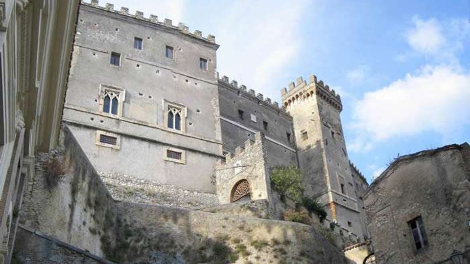 Veduta dal basso di una fortezza in pietra su una collina, con alte mura e torri merlate e piccole finestre. In primo piano si vedono i muri di contenimento e un edificio in pietra, con cielo azzurro sullo sfondo.