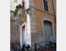 Angolo di strada in un centro storico con edificio intonacato color ocra e pietra bugnata, finestre con persiane e una piccola balconata con piante. Al piano terra si vedono porte e vetrine con grate, muri con graffiti e alcune biciclette e scooter parcheggiati lungo i paletti.