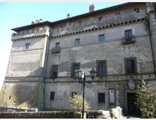 Facciata di un grande edificio storico in pietra e intonaco, simile a un castello o palazzo fortificato, con piccole finestre con inferriate, mensole sotto il tetto e un torrione laterale. In primo piano si vede un lampione a due lanterne e un piccolo spazio esterno con muretti e vegetazione.