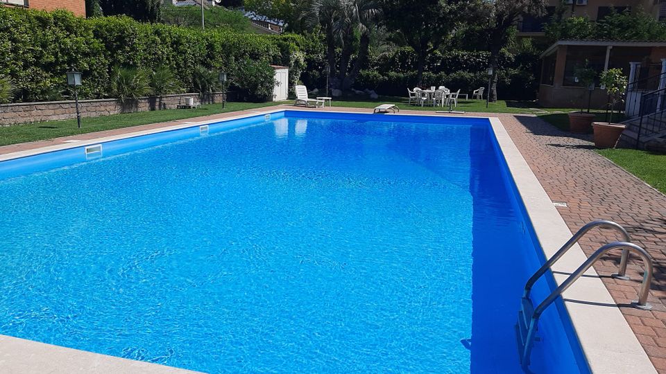 Piscina rettangolare all’aperto con acqua azzurra, bordata in pietra e circondata da pavimentazione in mattoni e prato. Sullo sfondo ci sono siepi e alberi, lettini e un tavolo con sedie vicino a edifici residenziali.