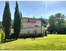 Ampio prato verde in un giardino di campagna con cipressi alti, cespugli e piccole palme lungo un muretto. Sullo sfondo si vede una casa a due piani con tetto in tegole e balcone, circondata da alberi e colline boscose.