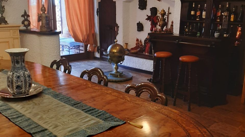 Sala da pranzo in stile tradizionale con grande tavolo ovale in legno intarsiato, lampadario e pavimento in cotto. Sullo sfondo si vedono un angolo bar con sgabelli, una vetrina con bottiglie e oggetti decorativi, e una porta-finestra con tende arancioni che conduce a un balcone.