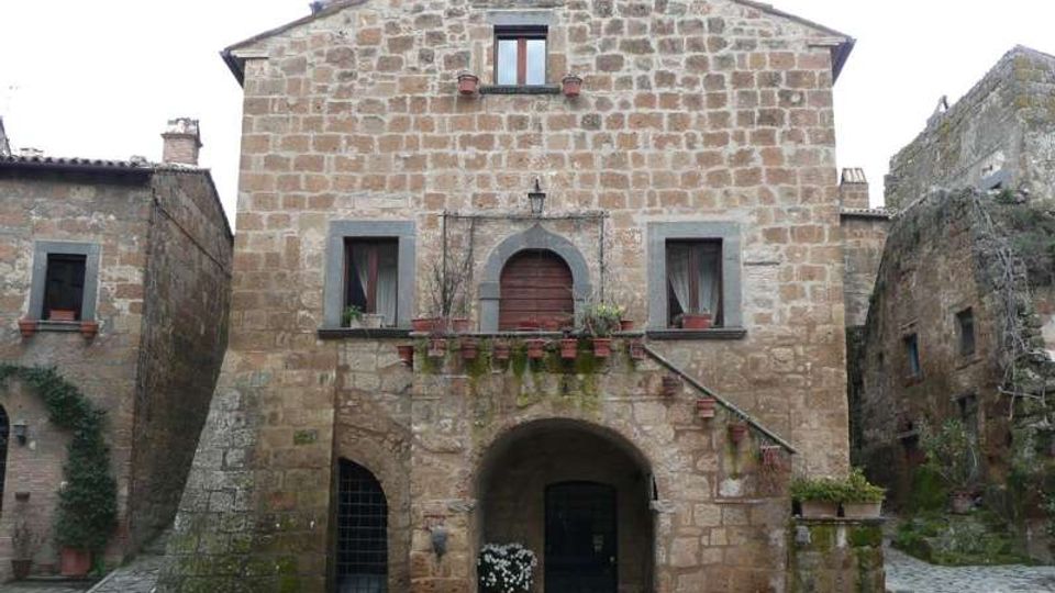 Facciata di una casa in pietra in un borgo storico, con grande arco d’ingresso al piano terra, finestre simmetriche e balconcino con porta ad arco e vasi di fiori. Pavimentazione in pietra e edifici adiacenti in muratura su entrambi i lati.