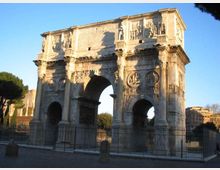 Arco monumentale romano in pietra con tre grandi aperture e ricchi bassorilievi decorativi sulle facciate. L’arco è circondato da una recinzione metallica, con alberi e edifici sullo sfondo sotto un cielo sereno.