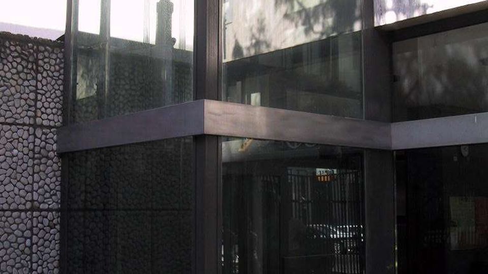 Angolo di un edificio moderno con struttura in metallo scuro e grandi pannelli di vetro, simile a un vano ascensore o ingresso vetrato. Accanto si vede un muro rivestito in pietre a mosaico e una pavimentazione esterna in pietra, con ombre di alberi riflesse sulle superfici.