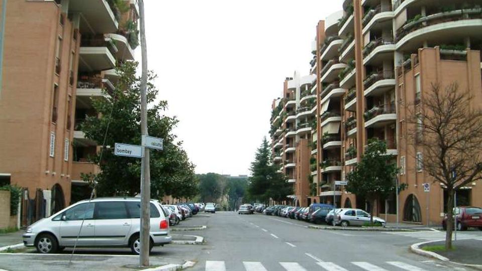 Strada residenziale ampia con attraversamento pedonale, auto parcheggiate ai lati e alberi lungo i marciapiedi. Su entrambi i lati si affacciano alti condomini in mattoni con balconi curvi e ringhiere bianche, con un palo segnaletico all’incrocio in primo piano.