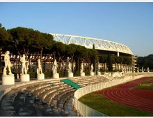 Stadio all’aperto con gradinate in pietra disposte a semicerchio, fiancheggiate da statue su piedistalli lungo il bordo superiore. In primo piano si vede una pista di atletica rossa e, sullo sfondo, una grande tribuna coperta con struttura metallica bianca e alberi alti dietro le gradinate.