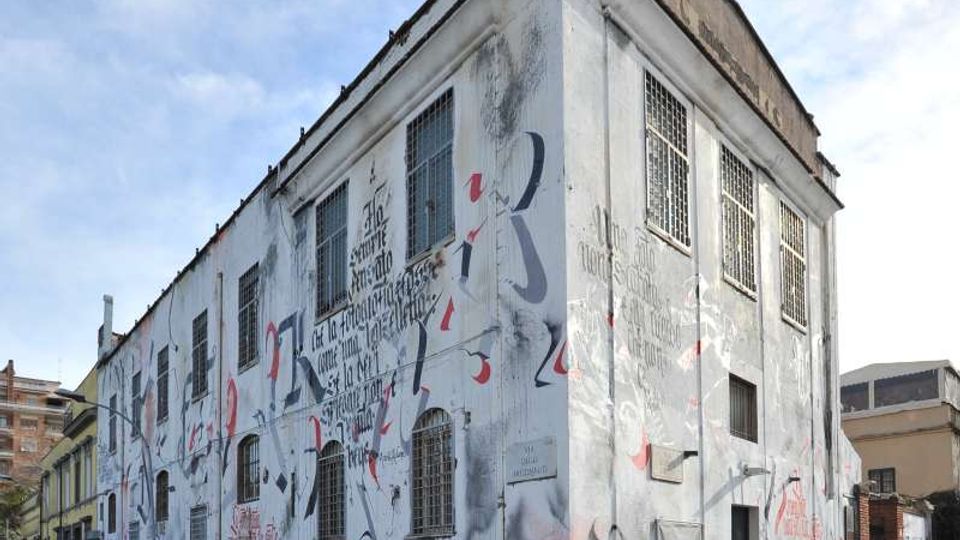 Edificio industriale a due piani con facciate bianche coperte da murales e graffiti, grandi finestre con griglie metalliche e spigolo su un incrocio stradale. Il contesto è urbano, con marciapiede, dissuasori lungo la strada e altri edifici sullo sfondo.