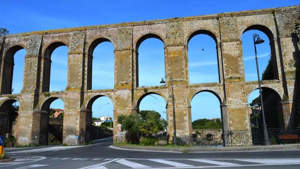 Antico acquedotto in pietra con due ordini di grandi arcate, che attraversa una strada asfaltata con segnaletica orizzontale. Sullo sfondo si vedono lampioni, una ringhiera e alberi sotto un cielo sereno.