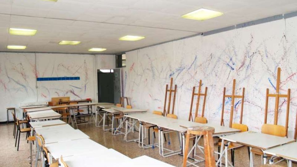 Aula di laboratorio artistico con tavoli bianchi disposti in file e sgabelli, illuminata da pannelli luminosi a soffitto. Lungo la parete destra ci sono cavalletti in legno e pareti bianche macchiate di schizzi di colore.