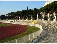 Gradinate in pietra di uno stadio all’aperto con pista di atletica rossa e recinzione metallica lungo il bordo. Sul coronamento delle gradinate si allinea una serie di statue bianche su piedistalli, con alberi alti sullo sfondo e una struttura coperta sulla destra.