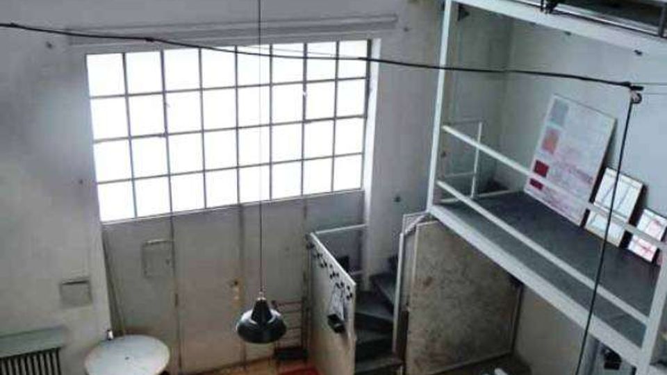 Interno di un loft a doppia altezza con grandi finestre ad arco e rettangolari a griglia che illuminano un ampio spazio. Sulla destra ci sono una struttura soppalcata in metallo con scala e parapetti, lampade a sospensione e arredi essenziali su pavimento piastrellato.