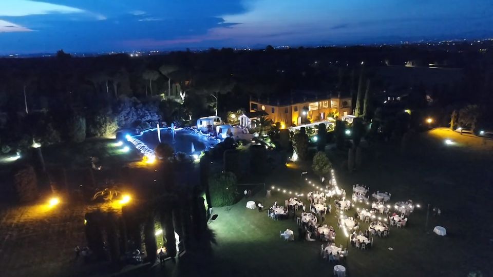 Veduta aerea notturna di una villa con giardini alberati, piscina illuminata e aree esterne con tavoli e luci decorative. Sullo sfondo si estende un paesaggio boscoso con luci urbane all’orizzonte sotto un cielo al crepuscolo.