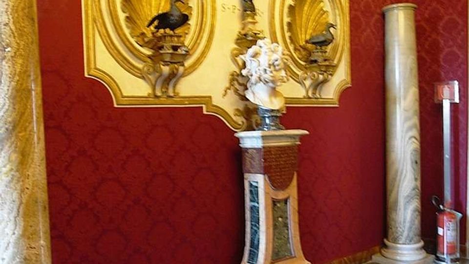 Interno di un edificio storico con pareti rosse damascate e pavimento in marmo a motivi geometrici. Sulla parete è fissata una grande targa decorativa dorata con nicchie a conchiglia e scritte, affiancata da due colonne in marmo e da un piedistallo con scultura.