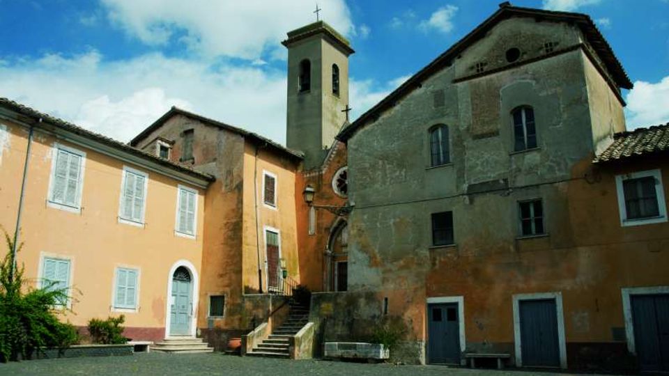 Piccola piazza lastricata con edifici in intonaco ocra e grigio e una chiesa affiancata da un campanile quadrato con croce in cima. Si vedono scale in pietra che salgono all’ingresso e facciate con finestre e porte sotto un cielo azzurro con nuvole.