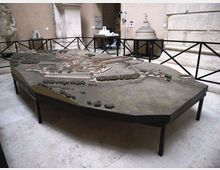 Grande plastico architettonico di una città antica esposto su un tavolo poligonale, circondato da barriere nere. L’ambiente è una sala museale con pareti in pietra e archi, con elementi scultorei sullo sfondo.