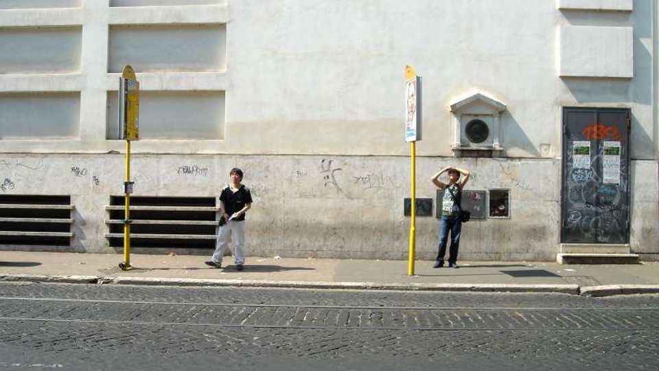 Marciapiede lungo una strada asfaltata davanti a un grande edificio con facciata chiara, riquadri rettangolari in rilievo e segni di graffiti nella parte bassa. Sono visibili due paline gialle di fermata, una griglia di aerazione e una porta scura con adesivi e scritte.