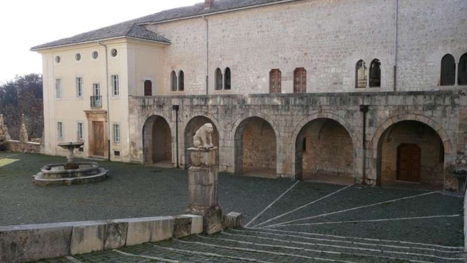 Cortile in pietra di un complesso storico con lunga facciata in muratura chiara e portico ad archi, affacciato su una piazza lastricata con scalinata. In primo piano si vede una piccola fontana circolare e un pilastro in pietra, con edifici a tetto in coppi sullo sfondo.