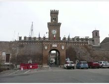 Porta cittadina fortificata in mattoni con grande arco centrale e torre merlata con orologio sulla facciata. Davanti c’è una piccola area stradale con auto parcheggiate e transenne, sotto un cielo coperto.