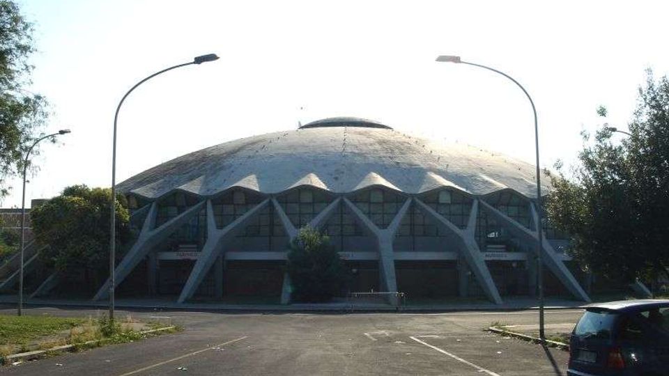 Edificio circolare con grande cupola ribassata e facciata scandita da archi e pilastri triangolari, con vetrate lungo il perimetro. Davanti si vede un ampio parcheggio asfaltato con lampioni e alberi ai lati.