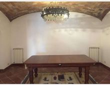 Interno di una sala con soffitto a volta in mattoni e pareti bianche, illuminata da un lampadario in ferro battuto. Al centro c’è un grande tavolo in legno sopra un tappeto, con pavimento in cotto e due radiatori ai lati.
