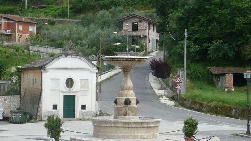 Piazza di un piccolo paese con una fontana in pietra al centro, su basamento circolare con catene e vasi. Sullo sfondo si vede una cappella bianca con porta verde, una strada che sale tra case sparse e un pendio collinare coperto di alberi.