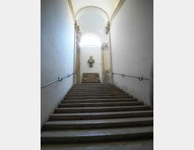 Scalinata interna in pietra con corrimano laterali, che conduce a un pianerottolo illuminato da una finestra ad arco sotto una volta. Sulla parete di fondo sono visibili un busto scultoreo e un bassorilievo decorativo tra elementi architettonici in pietra.