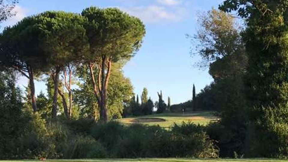 Campo da golf con ampio prato e area di tee in primo piano, circondato da vegetazione e alberi alti (inclusi pini a ombrello). Sullo sfondo si vede una leggera collina con cipressi e cielo sereno.