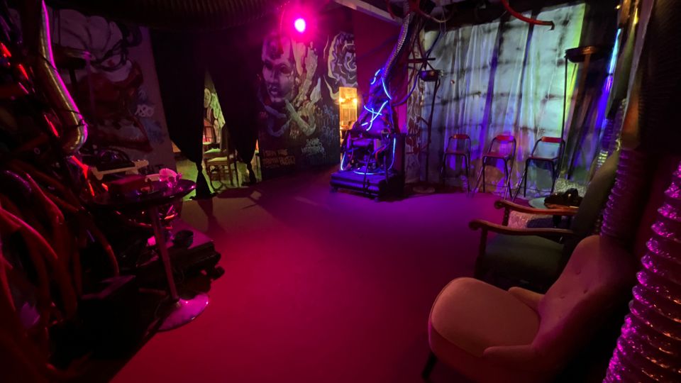 Interno di uno spazio artistico dall’aspetto industriale, illuminato da luci viola e rosa, con cavi e tubi a vista sul soffitto e murales sulle pareti. A destra ci sono tende chiare e sedie alte in fila, mentre in primo piano si vedono poltrone e arredi sparsi.