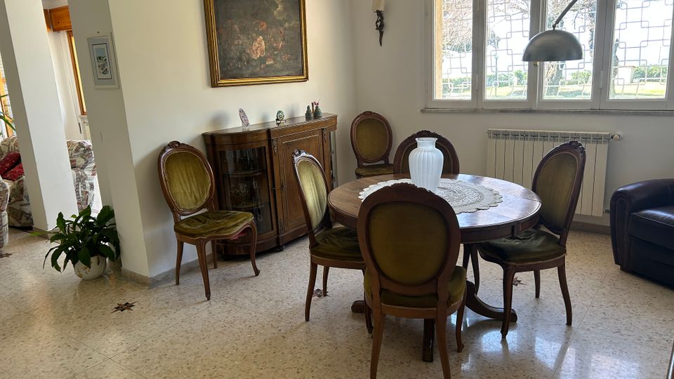 Sala da pranzo in un’abitazione, con tavolo rotondo in legno e sedie imbottite, credenza contro la parete e quadro incorniciato sopra. Ampie finestre con grata e radiatore sottostante illuminano un pavimento in graniglia; visibile anche una lampada ad arco e una poltrona in pelle.