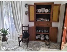 Angolo di soggiorno con pavimento in marmo e grande finestra con tende chiare che lascia entrare luce naturale. Accanto a una sedia in legno e a un vaso con pianta, c’è una credenza in legno con ante aperte che espone piatti e servizi da tè in ceramica.