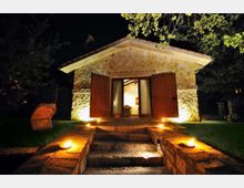 Piccolo edificio in pietra illuminato di notte, con tetto a due falde e grandi porte in legno aperte su un ingresso. Un breve vialetto con gradini in pietra e luci a terra conduce dalla zona erbosa fino alla soglia, circondato da alberi.